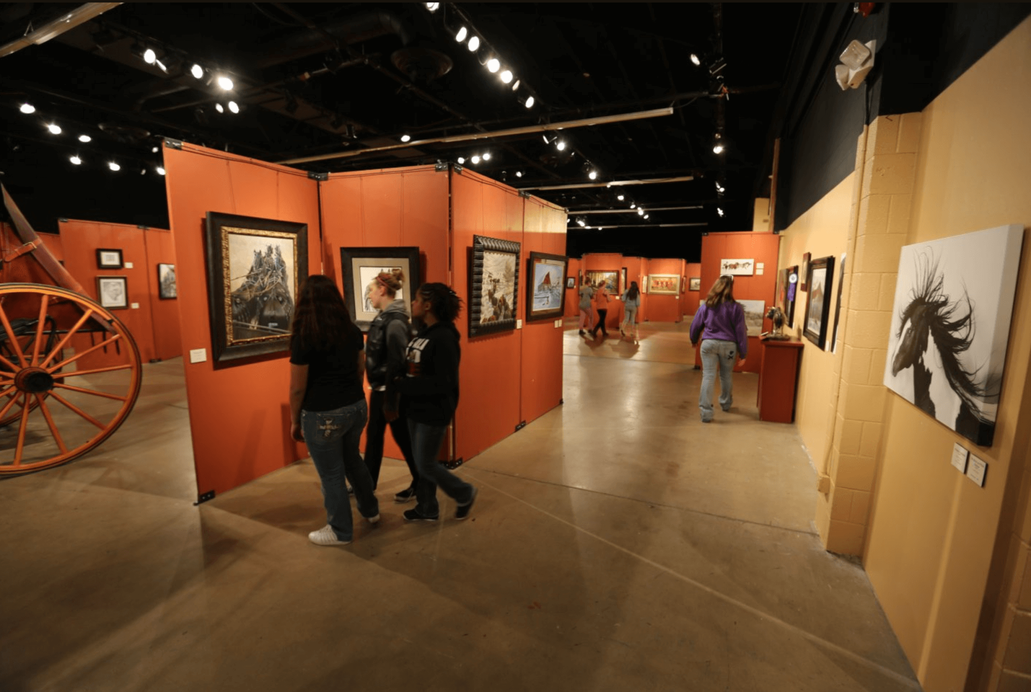 People stroll through a historic museum exhibit featuring a painting of a wild horse and a wagon wheel artifact.